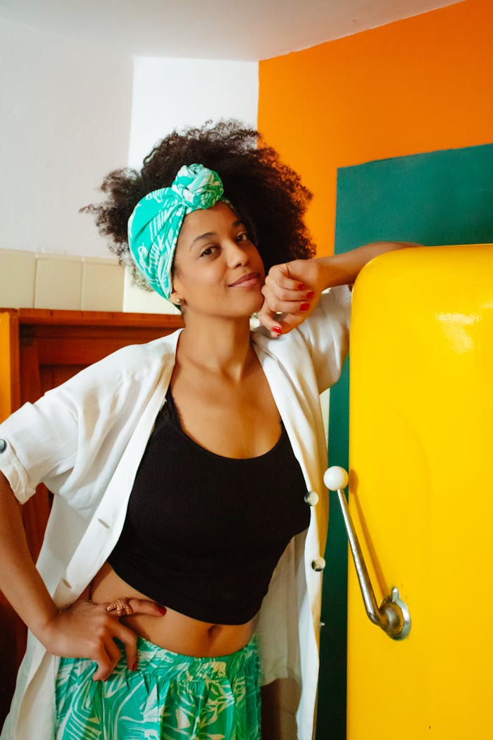 Woman with curly hair and headscarf smiling in a vibrant kitchen with a yellow fridge.